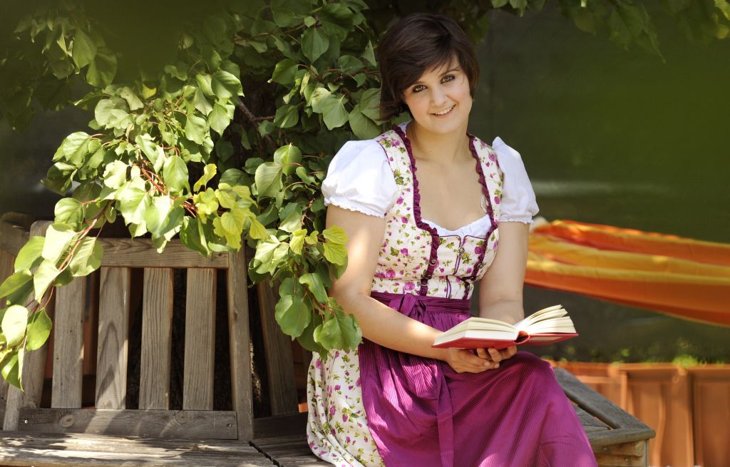 Woman in traditional dress reading a book on a wooden bench under a tree.