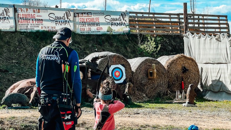 An adult and a child are standing on an archery course. In the background are targets and a sign with the words 'Parcours START'.