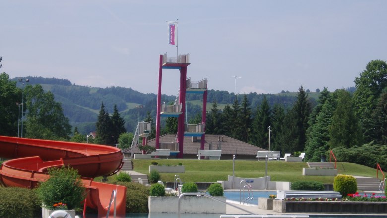 An outdoor pool with a slide, diving platform and green surroundings.