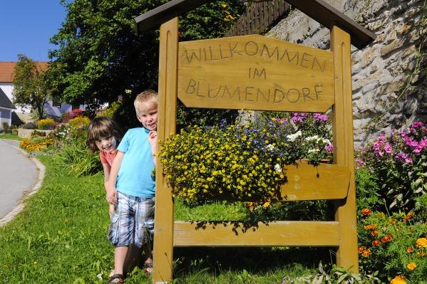 Two children stand next to a wooden sign with the inscription 'Welcome to the flower village', surrounded by colorful flowers.