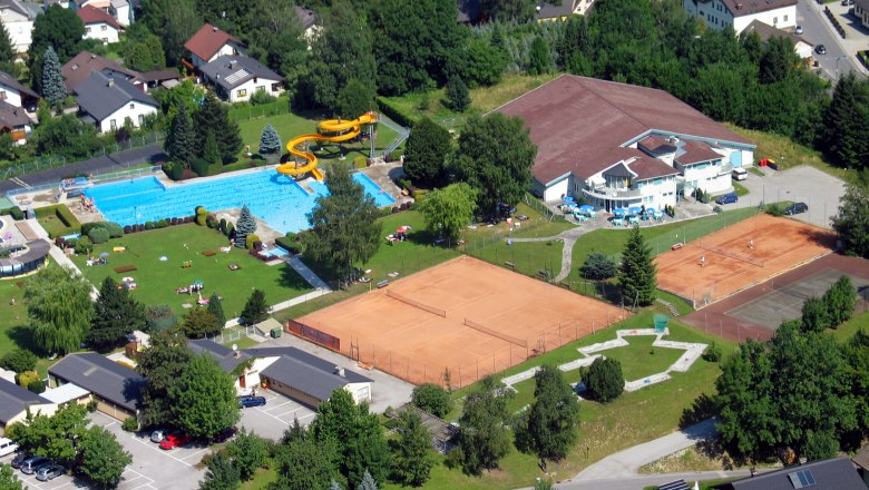 Aerial view of an outdoor pool with swimming pool, slide and tennis courts in a residential area.
