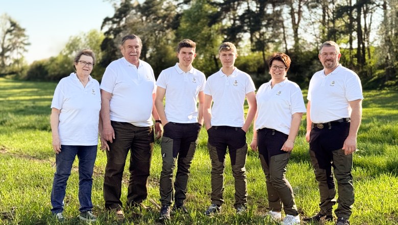 Six people in white polo shirts are standing on a meadow in front of trees.