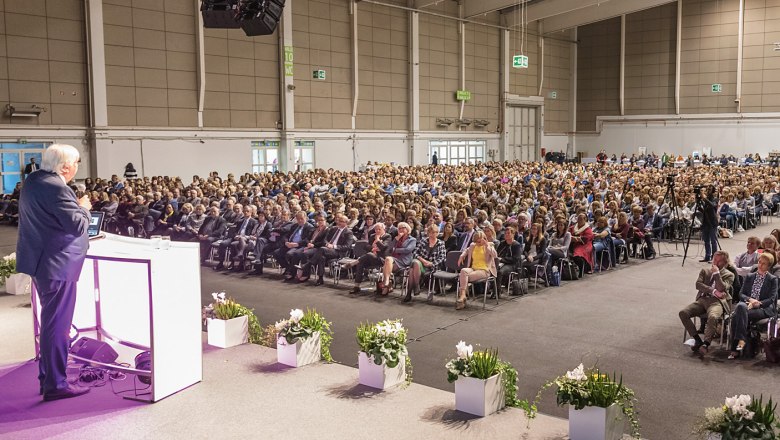 A speaker addresses a large audience in an exhibition hall.
