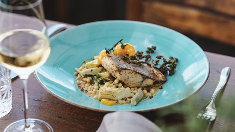 Gourmet dish with fish on a blue plate, glass of white wine next to it.