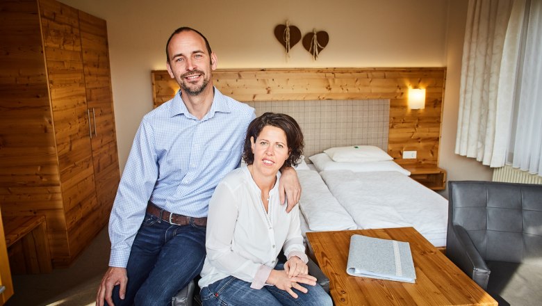 A couple is sitting in a cozy room with wooden furniture and a double bed.