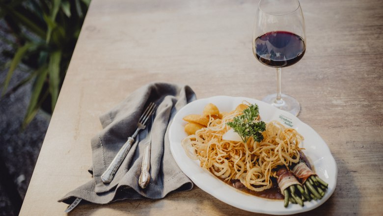 A plate of roast onions, potatoes and bacon beans, next to a glass of red wine on a wooden table.