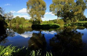 A calm river in the Dobersberg Nature Park with trees and a blue sky in the background.