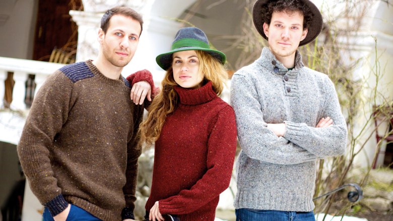 Three people in woolen sweaters and hats are standing outside a building.