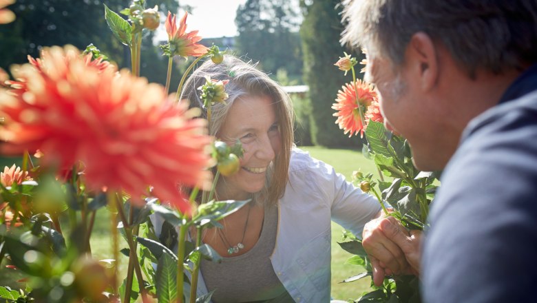 Two people smile at each other between blooming dahlias outdoors.