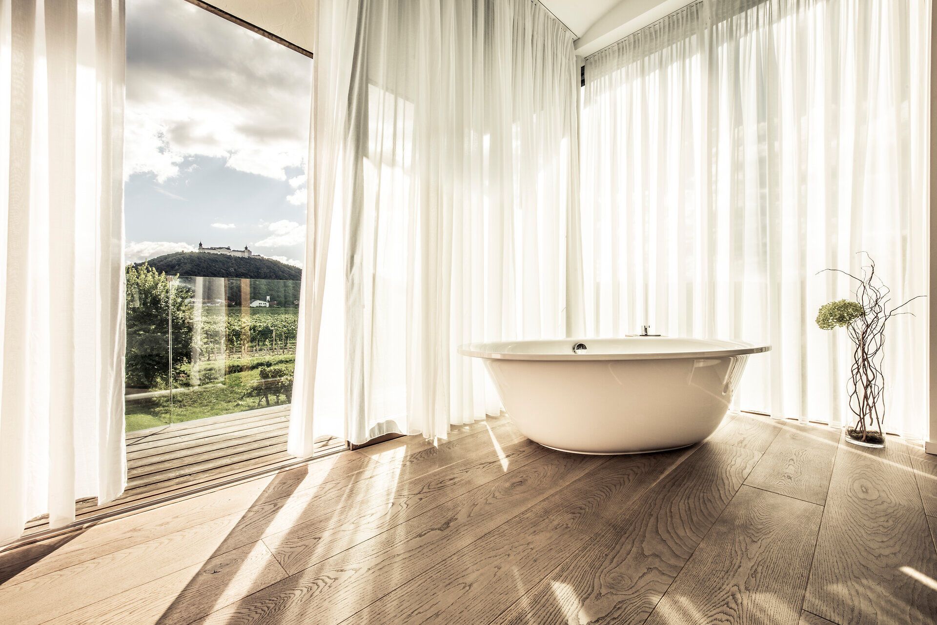 Einladend und hell strahlt der Raum mit seiner modernen Badewanne und den sanften Vorhängen, die das Licht filtern. Der Blick auf die sanften Hügel der Umgebung verspricht entspannende Stunden inmitten der Natur. Hier wird der Aufenthalt zu einem unvergesslichen Erlebnis der Ruhe und Erholung.
