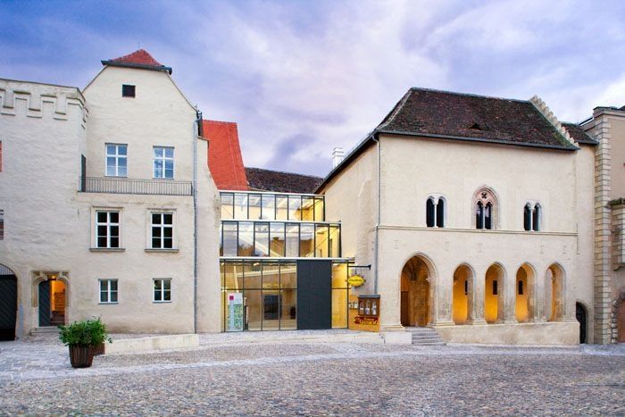 Historic building with a modern glass intermediate structure, cobblestones in the foreground.