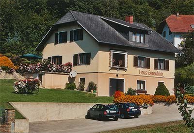 House Donaublick, © Hedi Kienberger A two-storey house with the inscription 'Haus Donaublick', surrounded by flowers and trees. Two cars are parked in front of the house.
