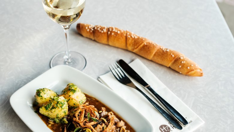 A table with a glass of white wine, a plate with a meat dish and dumplings, a long bread roll and cutlery on a napkin.