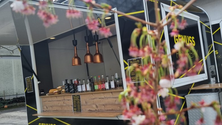 A black food truck with the inscription 'Genuss Fahrbrik' is parked outside. Blurred flowers can be seen in the foreground.