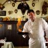 A chef in a white uniform flamb&eacute;es a dish in a rustic kitchen with hunting trophies on the wall.