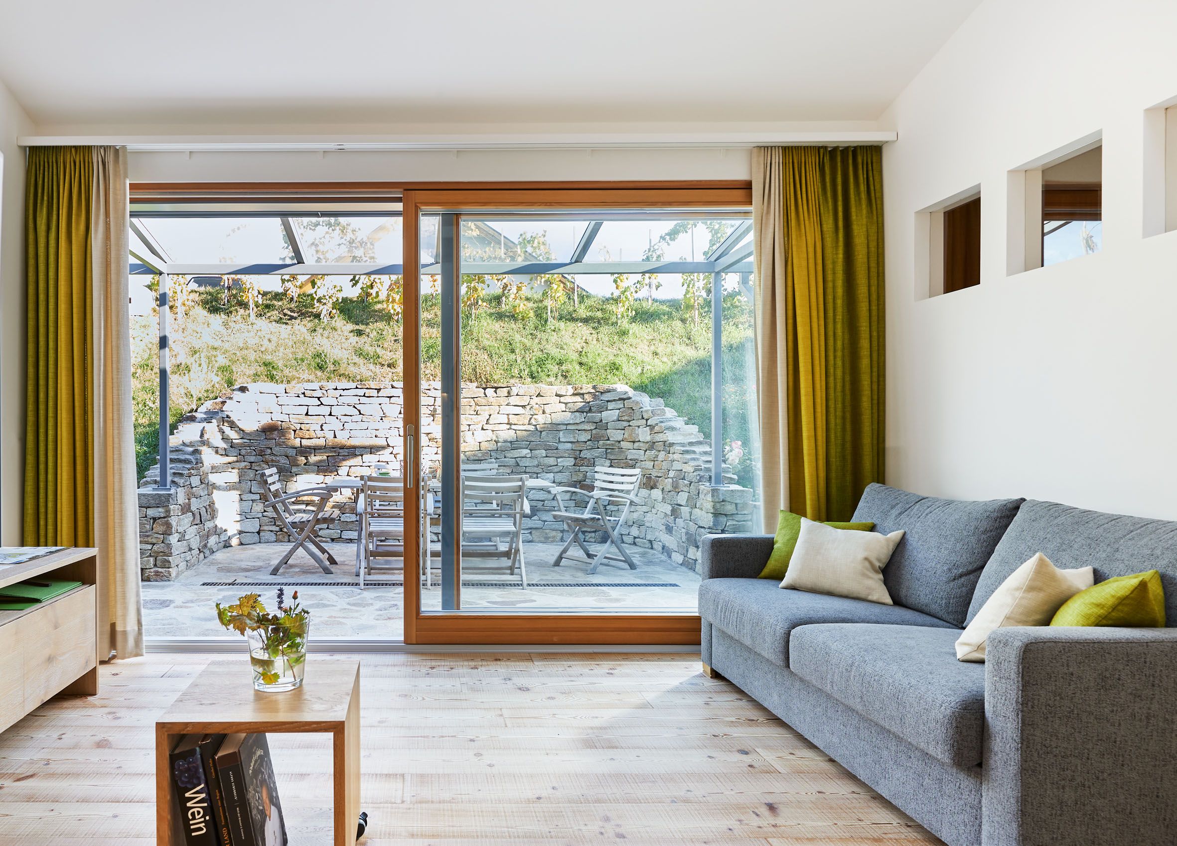 Interior view of a modern living room with gray sofa, wooden floor and large windows leading onto a terrace with stone wall and garden furniture.