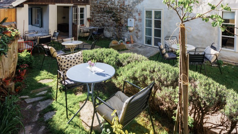 A cozy garden with tables and chairs, surrounded by plants and an old stone wall. Fairy lights hang over the garden.