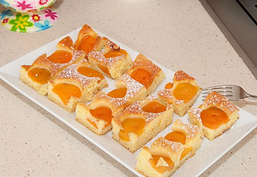 A rectangular tray with sliced apricot cake, sprinkled with powdered sugar, on a kitchen worktop.