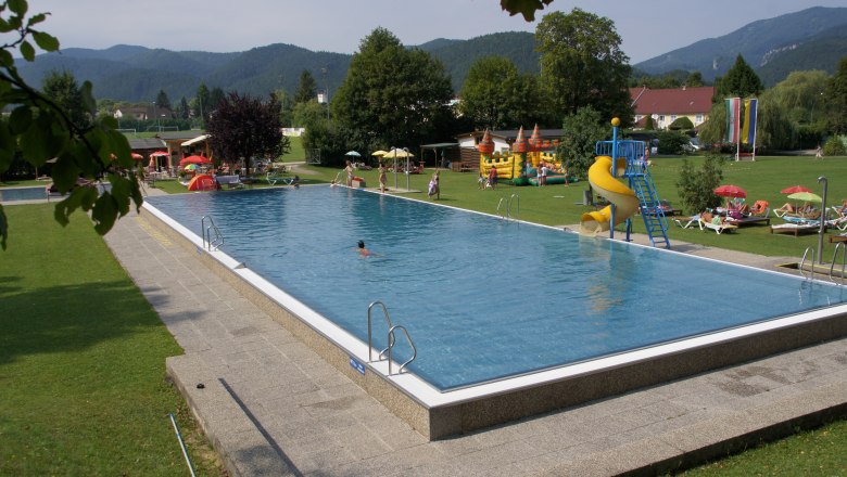 An outdoor pool with swimming pool, slide and sunbathing lawn, surrounded by trees and mountains.