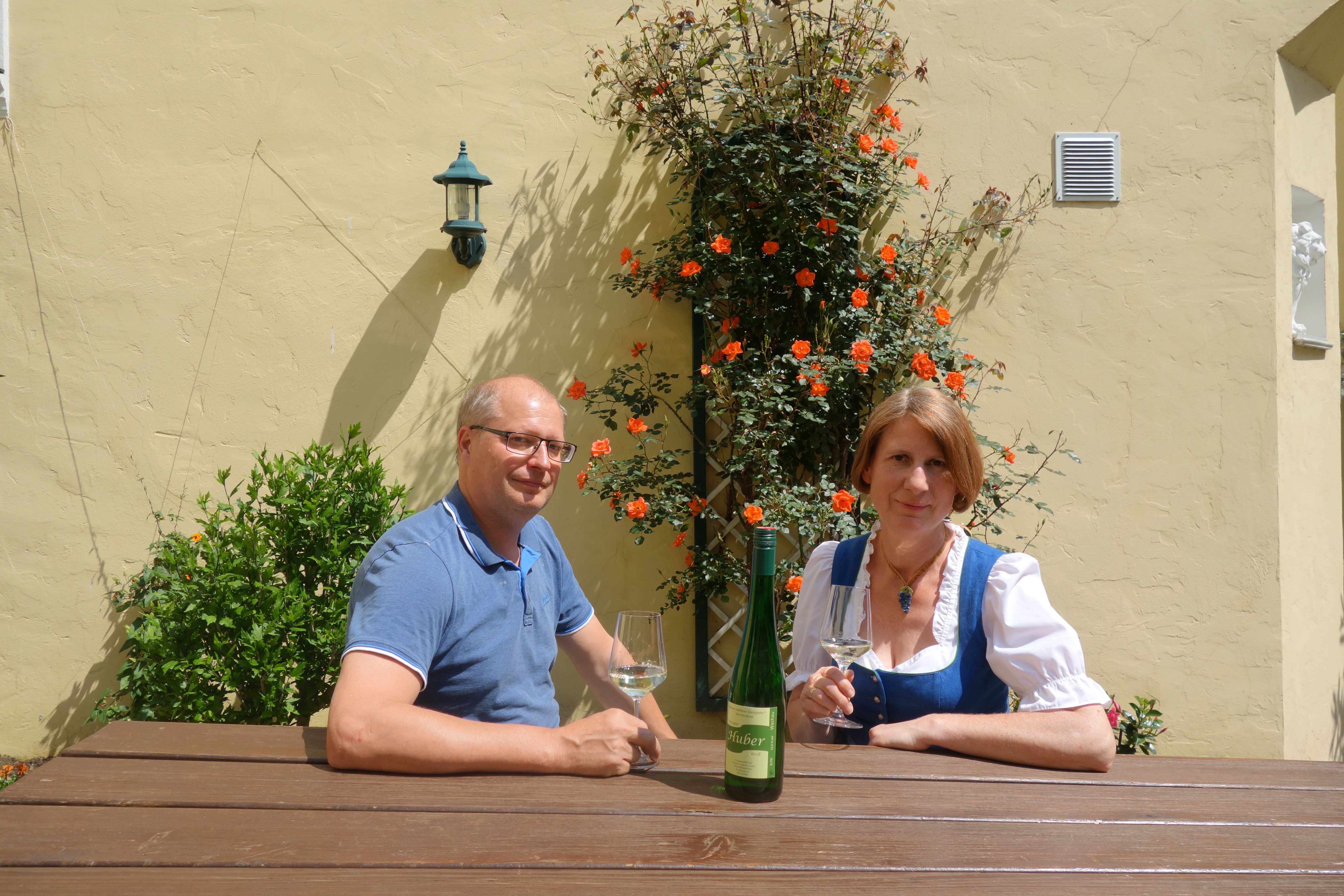 A man and a woman are sitting at a wooden table outside, with wine glasses and a bottle of wine. Orange flowers bloom in the background.