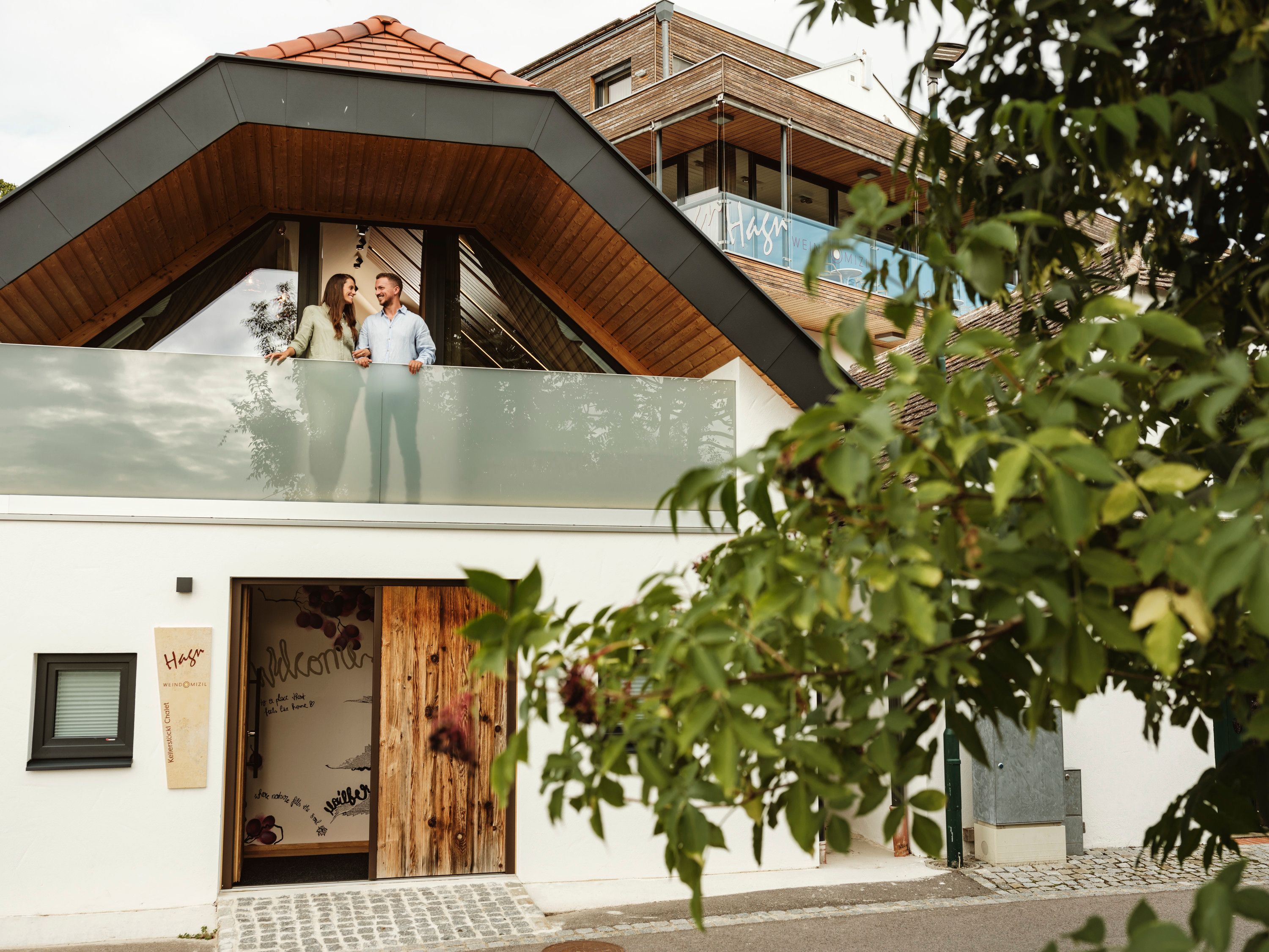 A modern building with a couple on the balcony, surrounded by trees.