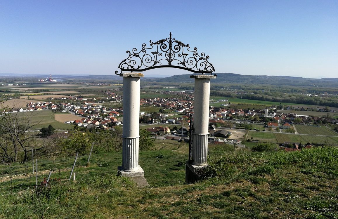 Two columns with an ornate arch on a hill overlooking a village and fields in the background.