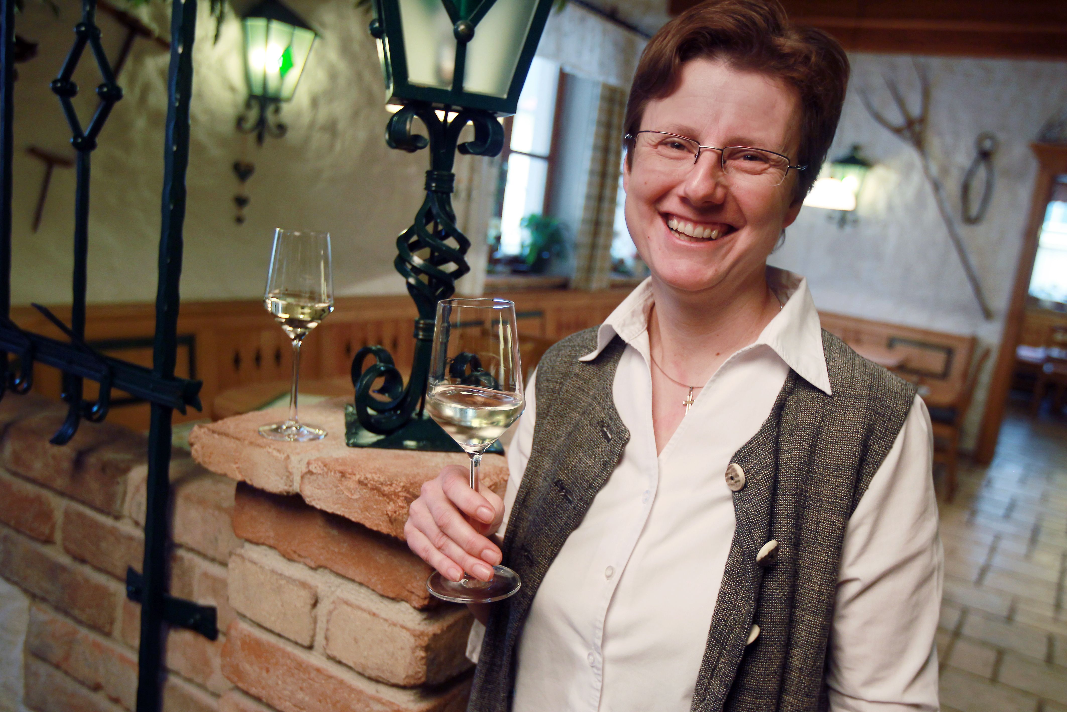 A smiling person holds a glass of white wine in a cozy room with a brick wall and a lantern.