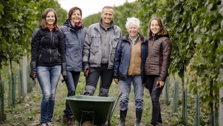 Five people stand smiling in a vineyard with a wheelbarrow.