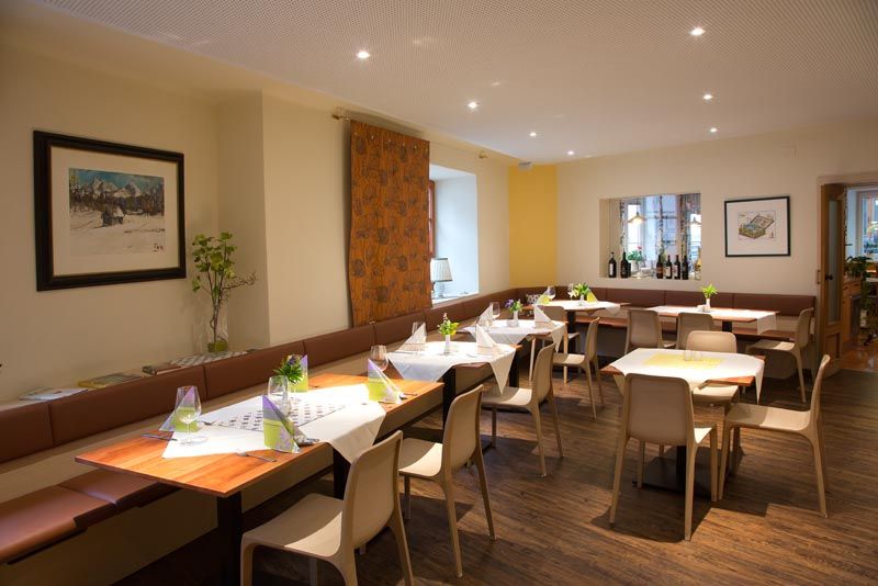 A modern dining room with laid tables, chairs and decorations. Pictures hang on the walls and there is a windowsill with bottles of wine.