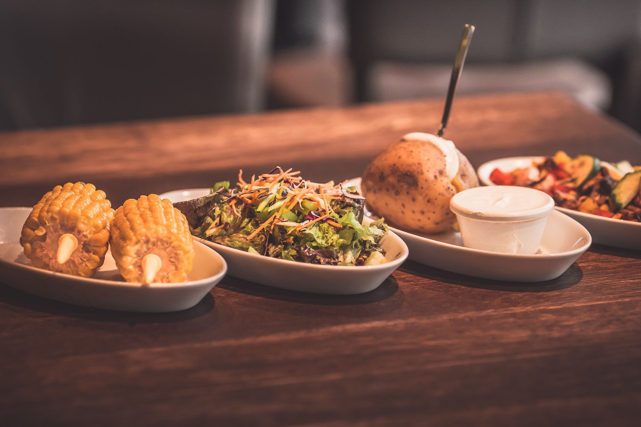 Various side dishes on a wooden table, including corn on the cob, salad, baked potato and dip.