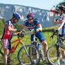 Three cyclists in sportswear, reading a map and drinking, against a mountain backdrop.