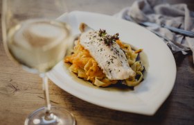 A plate of fish fillet on pasta and vegetables, with a glass of white wine next to it.