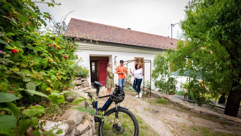 Three people are standing in front of a white house with a red roof, a bicycle in the foreground.
