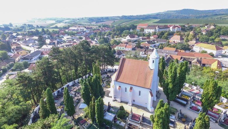 Aerial view of a church with a red roof, surrounded by a cemetery and trees, in a rural setting with houses and hills in the background.