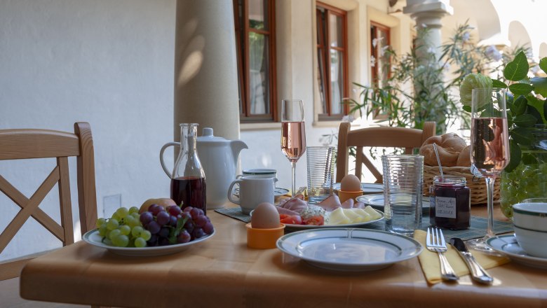 Breakfast table outside with grapes, ham, cheese, eggs, bread rolls and champagne glasses.