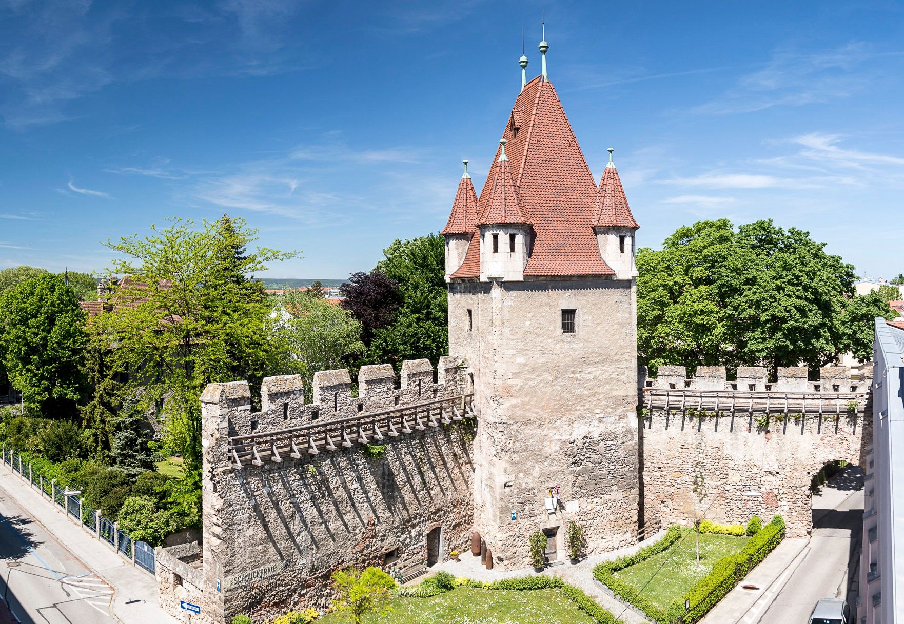 Rectangular tower with red roof and stone wall, surrounded by trees and roads.