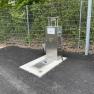 Metal water supply station next to a fence on asphalt.