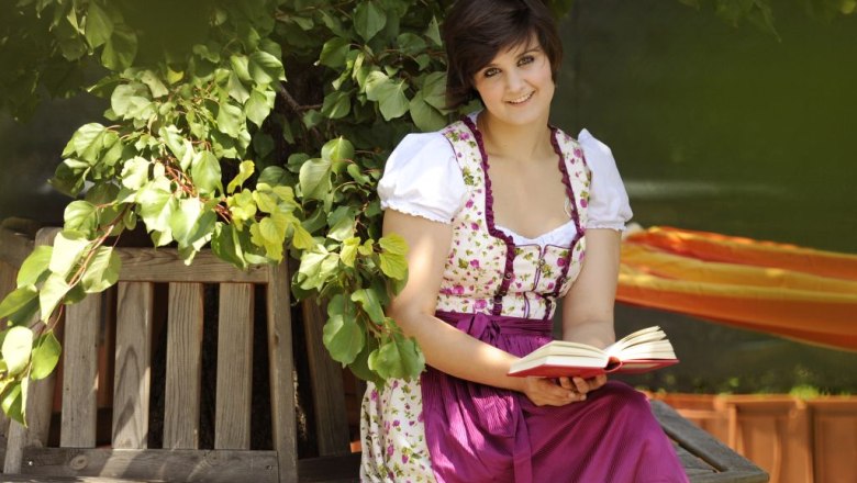 Woman in traditional dress reading a book on a wooden bench under a tree.