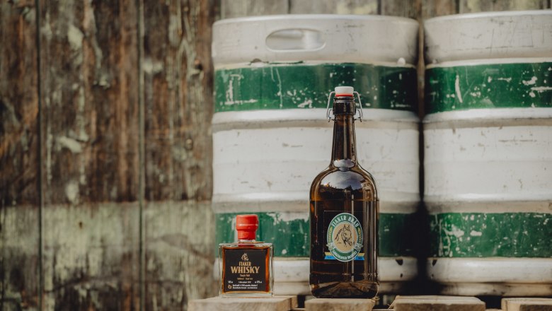 Bottle of Fiakerbräu and Fiaker Whisky in front of a wooden barrel.