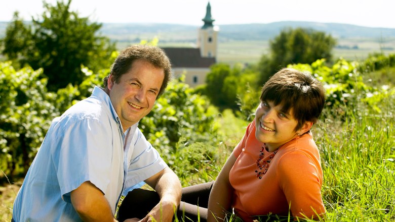 A man and a woman sit smiling on the grass in front of a church in a rural landscape.