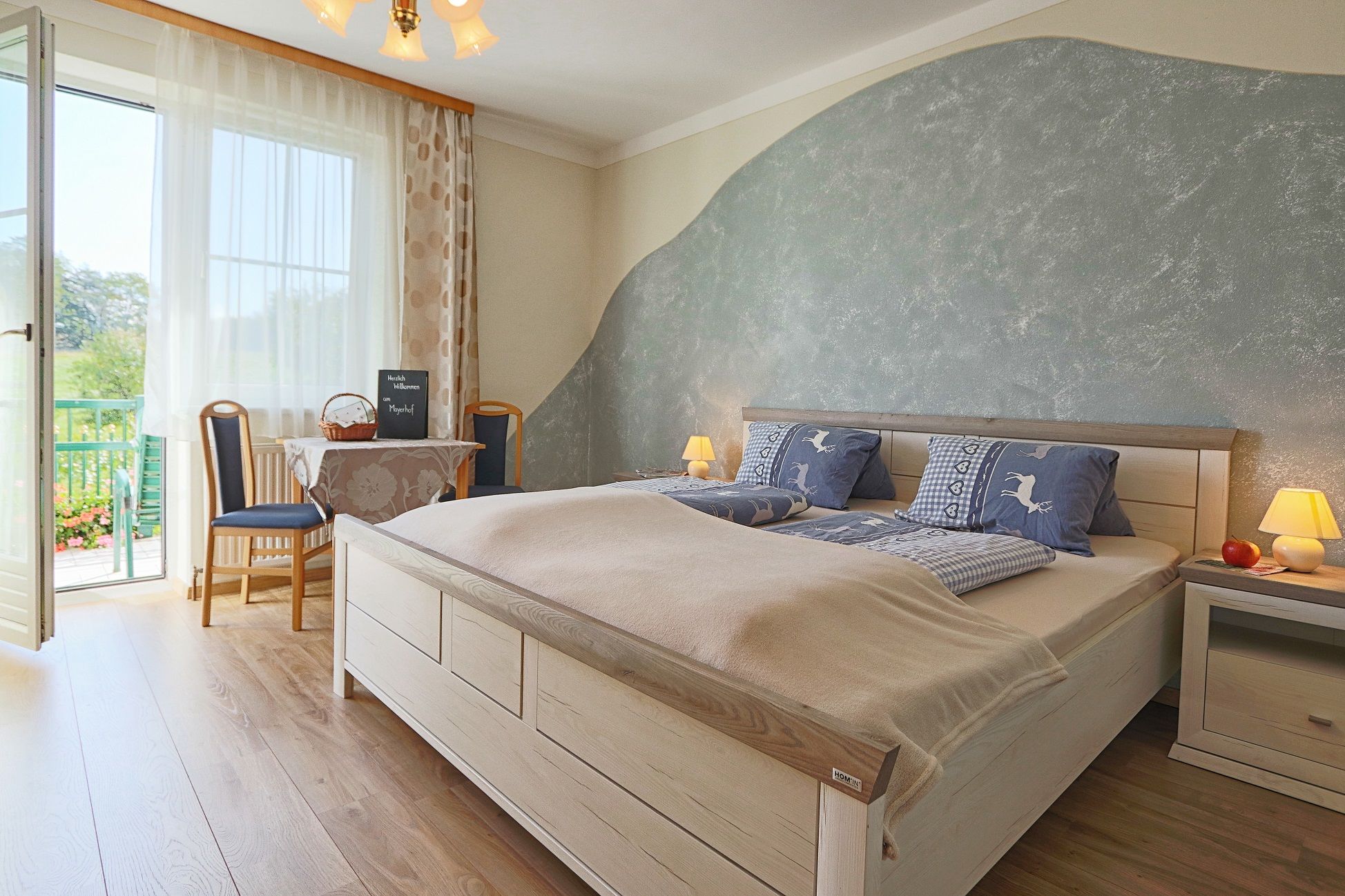 Cozy bedroom with double bed, blue bed linen, table with chairs and balcony access. Bright, inviting atmosphere.