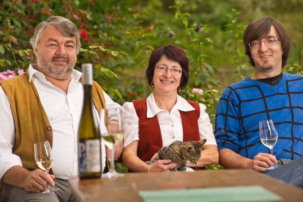 Three people sitting outside with glasses of wine and a cat.
