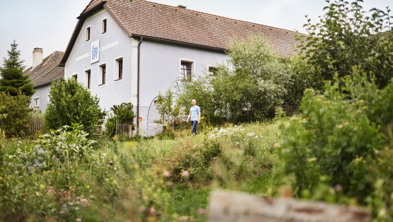 A man stands in front of a traditional Austrian house, surrounded by lush greenery and a garden.