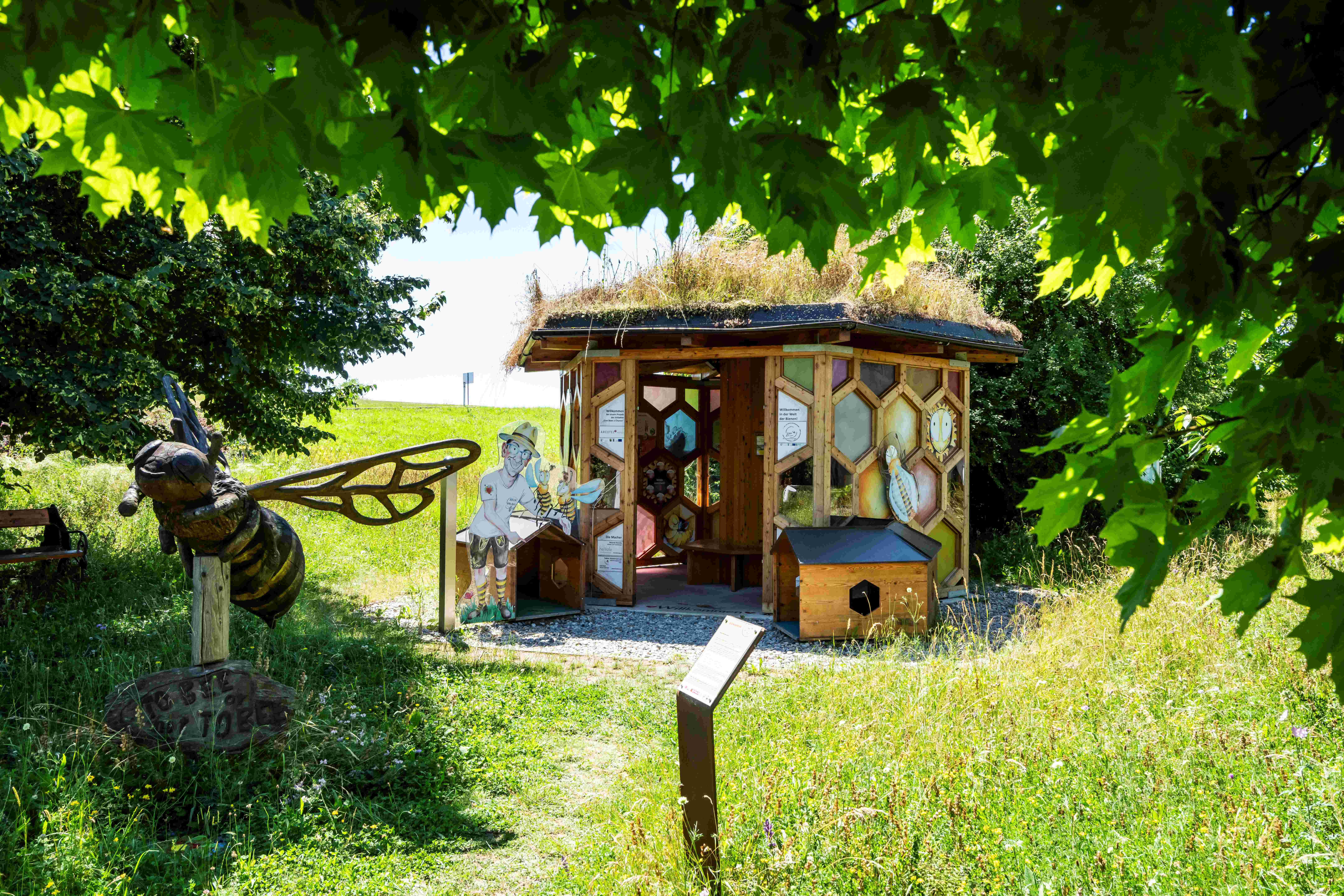 A bee pavilion with a grass roof and bee figures in the Klein-Pöchlarn Bee Park, surrounded by green nature.