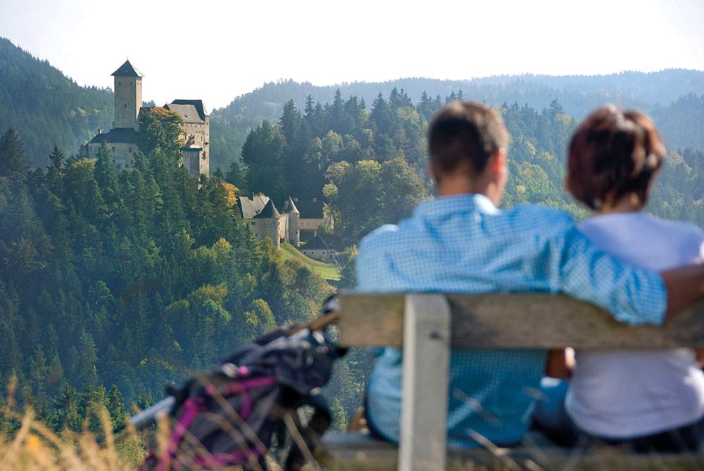 A couple sits on a bench with a view of a castle in a wooded landscape.