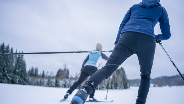 Two people cross-country skiing on a snow-covered trail, surrounded by trees.