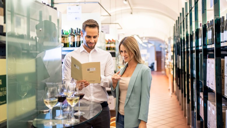 A man and a woman in a wine shop, the man is reading a wine list, the woman is smelling a glass of white wine.