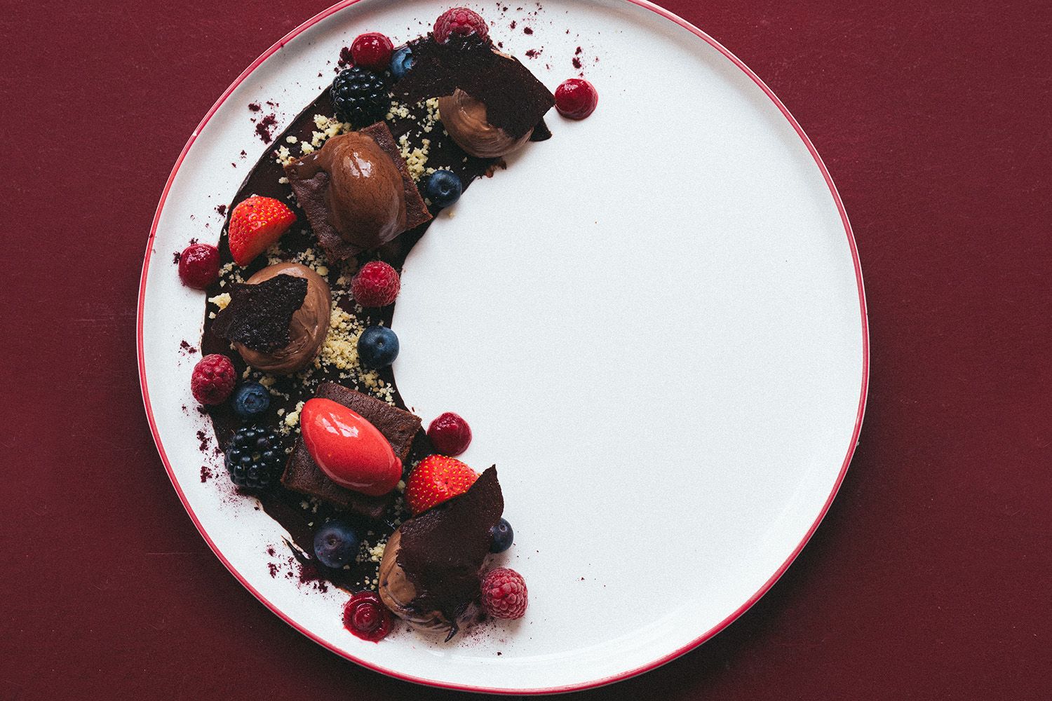 A dessert plate with chocolate mousse, berries and chocolate pieces on a red background.