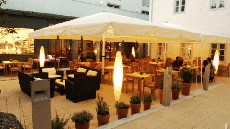 An illuminated guest garden with tables, chairs and plants in pots, surrounded by buildings.