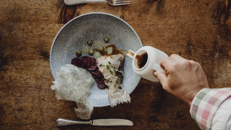 A plate of fish, beet and capers on a wooden table. A hand pours sauce from a jug over it.